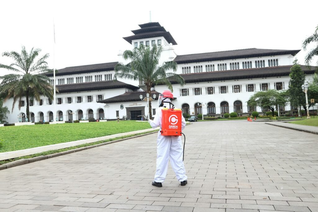 pexels-photo-16851355-16851355 A person in protective clothing disinfects an area in front of a historic building with palm trees.